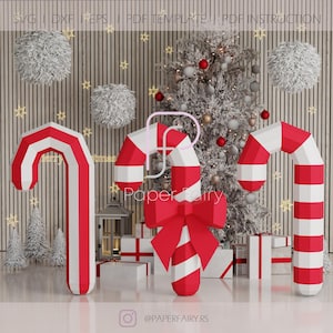 Peut inclure: Trois grandes cannes en papier rayées de rouge et de blanc avec un nœud rouge sur l'une d'elles. Les cannes sont placées devant un sapin de Noël avec des lumières blanches et des boules rouges et argentées. L'arrière-plan est un mur en bois avec des boules blanches et moelleuses suspendues au mur.