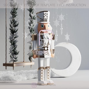 May include: White papercraft nutcracker figurine with a brown hat and gold accents. The nutcracker is standing on a white surface with a crescent moon and a swing set in the background.