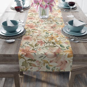 May include: A floral table runner with a cream background and a pattern of pink, peach, and yellow lilies and green leaves. The runner is set on a wooden table with place settings, wine glasses, and a vase of pink lilies.