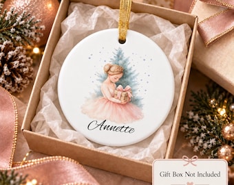 May include: A round white ceramic ornament featuring a watercolour illustration of a ballerina in a pink tutu holding a gift in front of a blue pine tree. The name Annette is printed in black script below the figure. A gold ribbon is attached.
