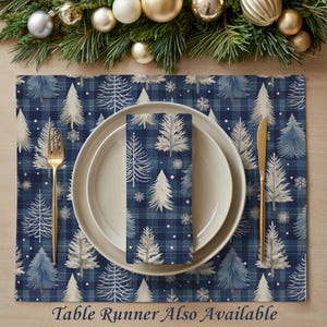 May include: A placemat and napkin set with a blue plaid pattern and white and blue Christmas trees. A gold fork and knife are on either side of a white plate. The text "Table Runner Also Available" is at the bottom.