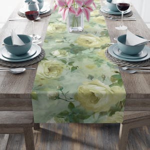 May include: A floral table runner featuring a watercolor design of large, pale yellow roses and green leaves on a soft green background. The runner is set on a wooden table with place settings, including teal bowls, plates, and wine glasses.
