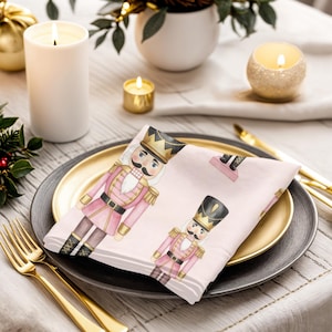 Peut inclure: Une serviette rose et blanche avec une illustration à l'aquarelle d'un soldat casse-noisette sur une assiette dorée et grise. La table est ornée de couverts dorés, de bougies et de verdure, créant un thème de vacances festif.