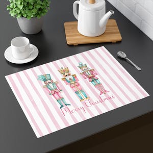 Op de afbeelding: Een rechthoekige roze en witte gestreepte placemat met drie aquarel notenkrakersoldaten in teal, roze en goud. De woorden "Merry Christmas" staan in cursief onderaan.