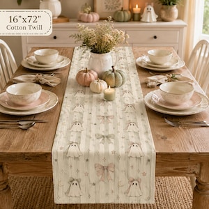 May include: A 16"x72" cotton twill table runner featuring a repeating pattern of white ghosts, pink bows, stars, and crescent moons on a light green and white striped background, centered on a wooden dining table with place settings.