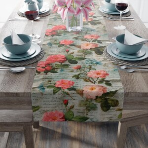 May include: A floral table runner featuring a vintage design with pink roses and script writing. The runner is placed on a wooden table set for a meal, with plates, bowls, and wine glasses.