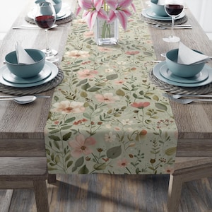 Op de afbeelding: Een bloemen tafelloper met een lichtgroene achtergrond en een patroon van roze en witte bloemen, groene bladeren en kleine rode accenten. De loper ligt op een houten tafel, gedekt met servies en wijnglazen.