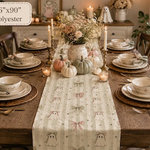 May include: A 16-inch by 90-inch polyester table runner featuring a repeating pattern of ghosts with bows, stars, and moons on a light green and white striped background. The runner is displayed on a wooden dining table set with place settings.