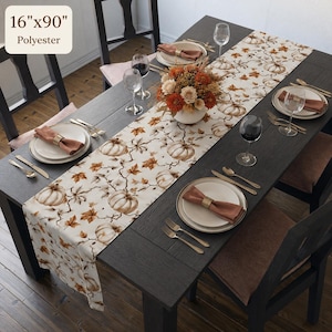 May include: A polyester table runner measuring 40.64 cm by 228.6 cm, featuring a repeating pattern of white pumpkins and brown autumn leaves on a cream background, laid across a dark wooden dining table set with plates, napkins, and glasses.