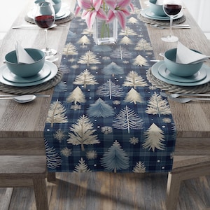 May include: A navy blue plaid table runner with a repeating pattern of stylized Christmas trees in white, light blue, and gold. The runner is set on a wooden table with place settings and a vase of pink flowers.