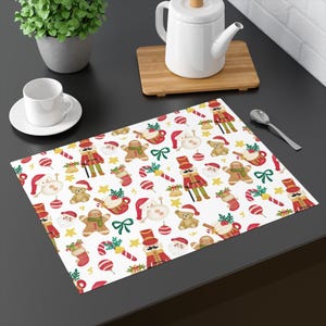 Op de afbeelding: Een wit kerst-thematisch placemat met een herhalend patroon van kerstmannen, peperkoekenmannen, notenkrakers, teddyberen en zuurstokken. De rechthoekige placemat is geschikt voor gebruik bij het eten of als decoratie.