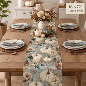 May include: A 16"x72" cotton twill table runner featuring a repeating pattern of white pumpkins, autumn flowers, and foliage on a light blue background. The runner is displayed on a wooden dining table set with plates, napkins, and a centerpiece.