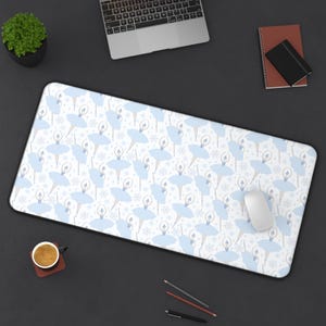 May include: A rectangular desk mat featuring a repeating pattern of light blue ballerinas and snowflakes on a white background. The mat has a black border and a white computer mouse rests on the surface. A cup of coffee, a laptop, and stationery are also visible.