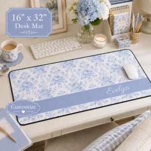 Può includere: Un tappetino da scrivania rettangolare di 40,6 cm per 81,3 cm, caratterizzato da un motivo floreale bianco e blu. Una fascia orizzontale azzurra attraversa la parte inferiore, riportando il nome Evelyn in corsivo bianco.