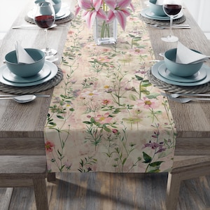 May include: A floral table runner with a watercolor design featuring pink and purple flowers and green foliage. The runner is placed on a wooden table set for a meal, with plates, bowls, and wine glasses.