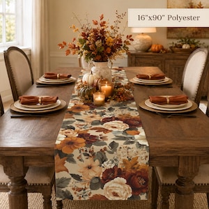Puede incluir: Un camino de mesa de poliéster de 40,6 cm x 228,6 cm con un estampado floral de flores y hojas en tonos crema, naranja y rojo oscuro. El camino está centrado sobre una mesa de madera con platos blancos, servilletas naranjas y servilleteros dorados.