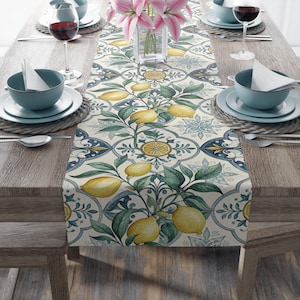 May include: A table runner with a lemon and leaf design. The runner features a cream-colored background with a repeating pattern of lemons, green leaves, and blue floral accents. The runner is placed on a wooden table set with dishes, glasses, and flowers.