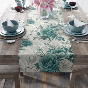 Op de afbeelding: Een bloemen tafelloper met grote teal rozen en witte duiven op een crèmekleurige achtergrond. De loper ligt op een houten tafel met couverts, wijnglazen en een vaas met roze lelies.