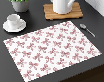 May include: White placemat featuring a repeating pattern of pink bows. A white teacup and saucer, a teapot, and a spoon are also visible on the dark surface. The bows are evenly spaced across the surface.