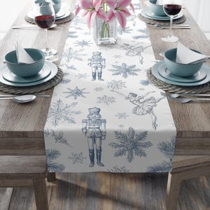 May include: A white table runner with a blue snowflake, ballerina, and nutcracker print. The runner is placed on a wooden table set for a meal, with plates, bowls, and wine glasses. Pink flowers are in a vase.