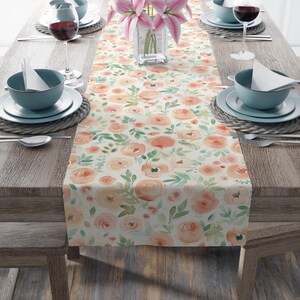 May include: A floral table runner featuring a watercolor design of peach and pink roses with green leaves on a cream background. The runner is placed on a wooden table set with tableware, including plates, bowls, and wine glasses, creating an elegant dining scene.