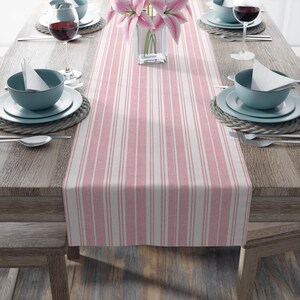 May include: A pink and white striped table runner is displayed on a wooden table, set for a meal. The table is set with plates, bowls, silverware, and wine glasses. A vase of pink flowers sits in the center.