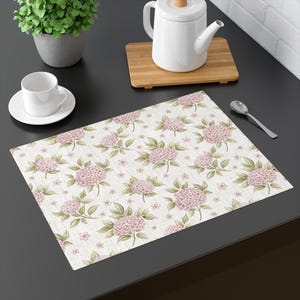 Puede incluir: Un mantel individual rectangular con un estampado floral de hortensias rosas y hojas verdes sobre un fondo crema. El mantel individual está sobre una superficie oscura, con una taza y platillo blancos, una tetera y una cuchara.