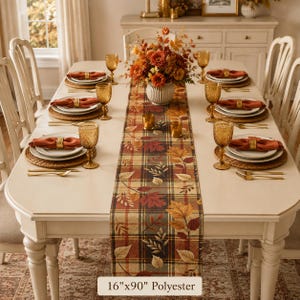 May include: A 16-inch by 90-inch polyester table runner featuring a plaid pattern with autumn leaves in shades of orange, red, and brown. The runner is displayed on a dining table set with white plates, orange napkins, gold napkin rings, and amber glassware.