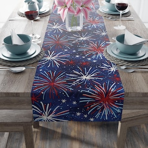 Puede incluir: Un camino de mesa rectangular con fondo azul oscuro y un patrón repetitivo de fuegos artificiales en rojo, blanco y azul claro, junto con pequeñas estrellas. La tela está extendida sobre una mesa de madera bajo un florero de cristal.