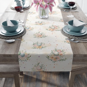 May include: A cream-colored table runner with a floral pattern featuring pastel-colored orbs and flowers. The runner is set on a wooden table with place settings, wine glasses, and a vase of pink lilies. The overall theme is elegant dining.