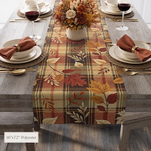 May include: A 16-inch by 72-inch polyester table runner featuring a plaid pattern in shades of tan, brown, and red, decorated with a print of autumn leaves. The runner is displayed on a wooden table with place settings and a floral centerpiece.
