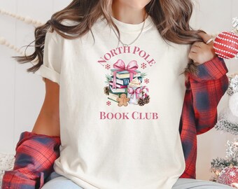 Könnte beinhalten: Cremefarbenes T-Shirt mit rosa Aufdruck „NORTH POLE BOOK CLUB“. Die Grafik zeigt einen Stapel Bücher, eine rosa Schleife, Schneeflocken, einen Lebkuchenmann und eine Zuckerstange.