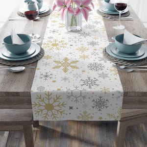 May include: A white table runner with a snowflake pattern in gold and gray. The runner is placed on a wooden table set for a meal, with plates, bowls, silverware, and wine glasses. A vase of pink flowers is in the center.