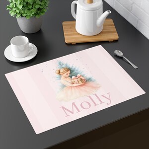 Op de afbeelding: Een roze placemat met verticale strepen toont een aquarel illustratie van een ballerina die een cadeau vasthoudt. De naam "Molly" is in een zachtroze lettertype eronder gedrukt. Een kleine kerstboom staat achter de ballerina.