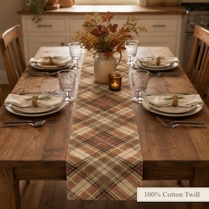 Puede incluir: Una mesa de comedor de madera con un camino de mesa a cuadros en tonos marrón, beige y crema. La mesa cuenta con cuatro cubiertos con platos blancos, servilletas plegadas con anillos de madera, vasos texturizados y un jarrón central con follaje.