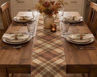 May include: A 100% cotton twill table runner featuring a brown, tan, and cream plaid pattern. The runner is centered on a wooden dining table set with white plates, folded napkins with rosemary sprigs, clear textured glasses, and a floral centerpiece.