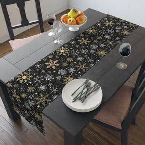 May include: A black table runner with a gold and silver snowflake pattern. The runner is on a dark wooden table set with plates, silverware, wine glasses, and a bowl of apples. The table is set for a holiday meal.