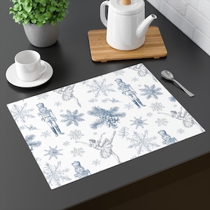 Op de afbeelding: Witte rechthoekige placemat met een blauw toile-de-jouy patroon met sneeuwvlokken, notenkrakers en ballerina's. De placemat ligt op een donkere ondergrond, met een theepot en een kopje op de achtergrond. Een decoratief item voor de eettafel.