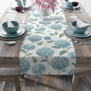 May include: A white table runner featuring a watercolor pattern of blue hydrangea flowers and green leaves. The runner is placed on a wooden table set for a meal, with plates, bowls, silverware, and wine glasses.