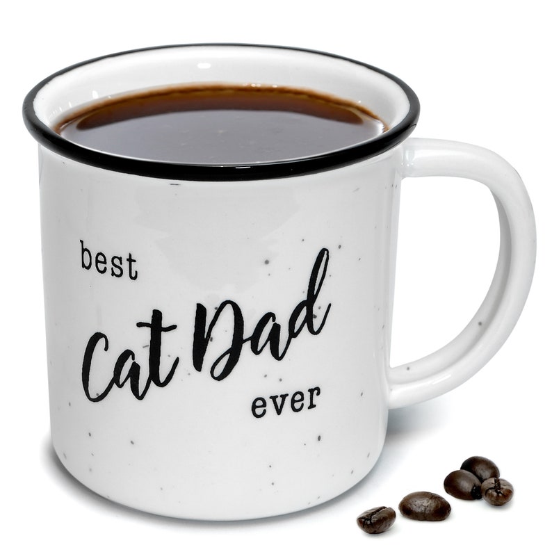 Pode incluir: Caneca de cer&acirc;mica branca com manchas pretas e uma borda preta. A caneca tem o texto "best cat dad ever" impresso em preto. A caneca est&aacute; cheia de caf&eacute; e h&aacute; gr&atilde;os de caf&eacute; espalhados sobre uma superf&iacute;cie branca.