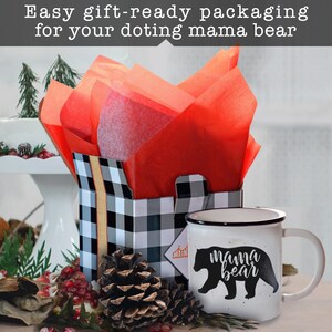 May include: A white ceramic mug with a black rim and handle. The mug has a black silhouette of a bear with the text "mama bear" printed on it. The mug is sitting on a table next to a black and white checkered gift box with red tissue paper inside. A pine cone is in front of the box.