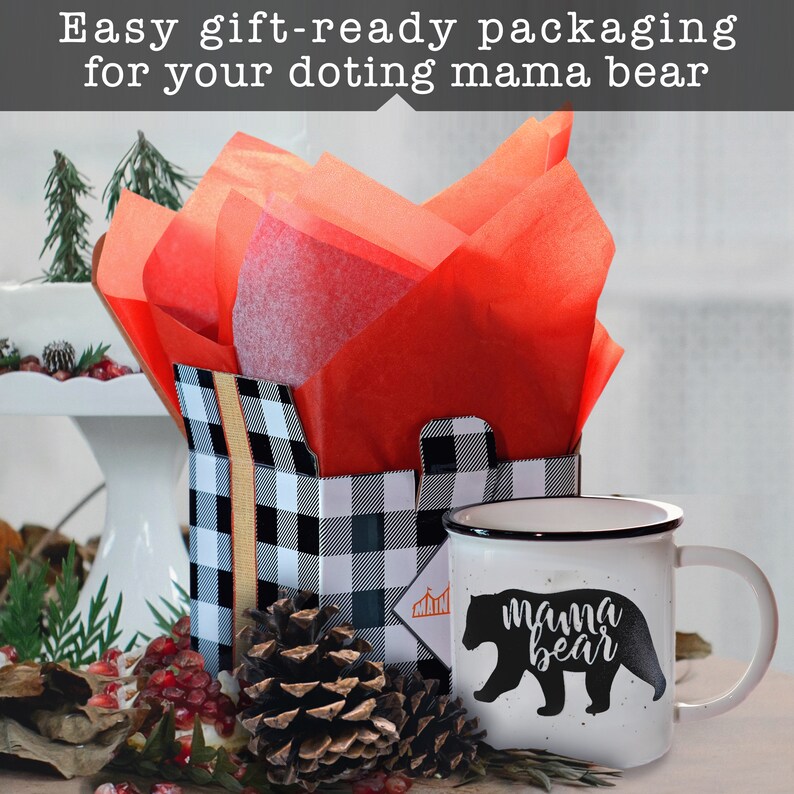May include: A white ceramic mug with a black rim and handle. The mug has a black silhouette of a bear with the text "mama bear" printed on it. The mug is sitting on a table next to a black and white checkered gift box with red tissue paper inside. A pine cone is in front of the box.
