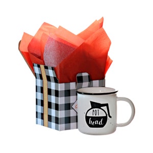 May include: A white ceramic mug with a black rim and the words "POT head" printed on it, next to a black and white checkered gift bag with red tissue paper. The mug has a coffee pot design.
