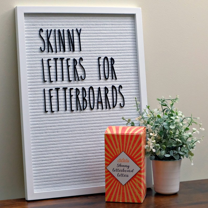 May include: A white letter board with the text "Skinny Letters for Letterboards" in black letters. A box of "Skinny Letterboard Letters" is in front of the letter board. The box is orange and yellow with a sunburst design. A small potted plant is to the right of the box.