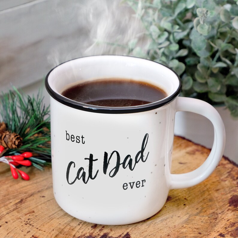 Pode incluir: Caneca de cer&acirc;mica branca com texto preto que diz "best cat dad ever". A caneca est&aacute; cheia de caf&eacute; e vapor est&aacute; subindo do topo.