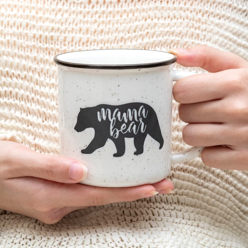 May include: White ceramic mug with a black silhouette of a bear and the text "mama bear".
