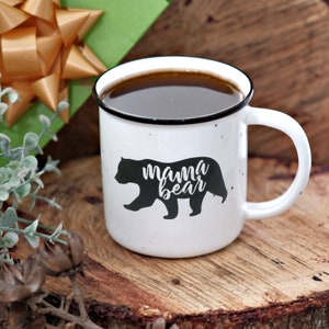 May include: White ceramic mug with a black bear silhouette and the text "mama bear" in black lettering. The mug is filled with dark brown liquid.