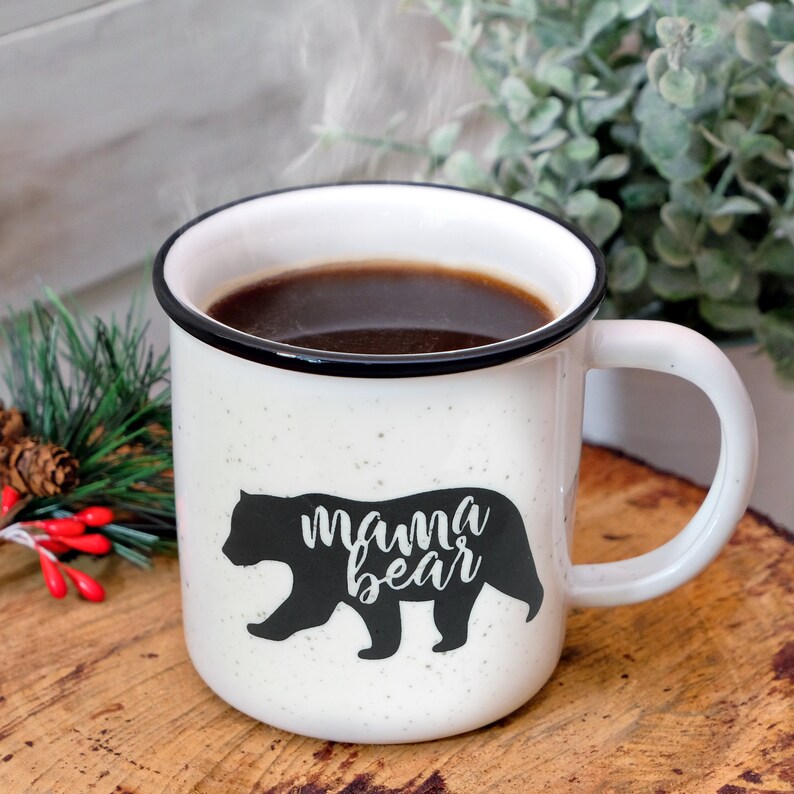May include: White ceramic mug with a black rim and a black silhouette of a bear with the text "mama bear" on it. The mug is filled with coffee and steam is rising from the mug.