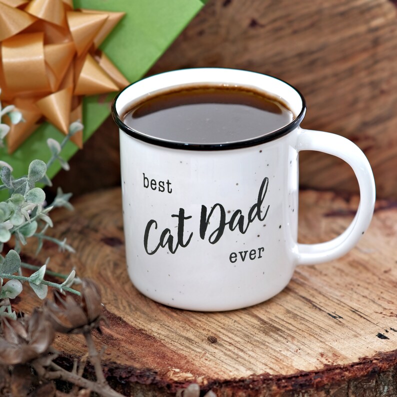 Pode incluir: Caneca de cer&acirc;mica branca com borda e al&ccedil;a pretas. A caneca tem manchas pretas e diz "best Cat Dad ever" em letras pretas.