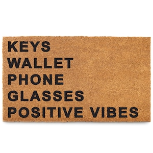 May include: A rectangular doormat made of natural brown coir fiber. The mat features the words "KEYS WALLET PHONE GLASSES POSITIVE VIBES" in bold, black capital letters. The mat is designed for placement at an entryway.
